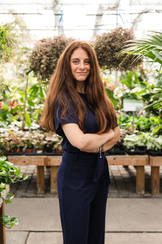 Me'Shelle Bishop standing in a tailored navy blue suit with short sleeves and long, beautiful, thick auburn hair that hangs over her shoulders and part way down the middle part of her upper arm. She is smiling and has her arms folded, appearing confident. She is standing in a lovely green house with many beautiful flowers in hanging post and sitting on wooden stands. There are about 100 flowers in the scene.